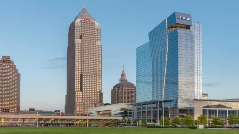 Cleveland cityscape showcasing the Hilton Cleveland Downtown, a modern glass-clad building, with adjacent skyscrapers and lush landscaping.