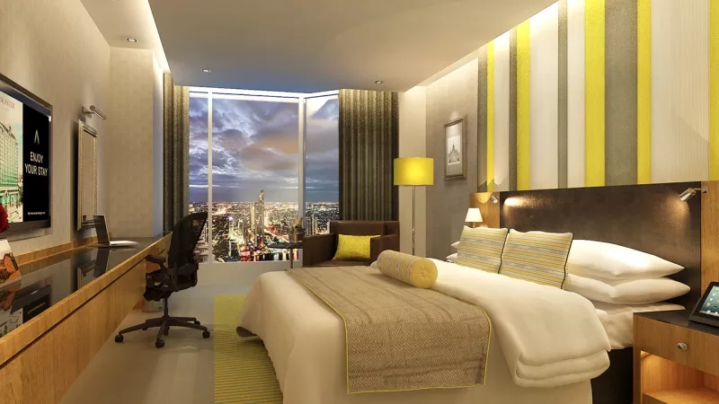 Lancaster Bangkok Hotel's deluxe room has a bed, desk, chair, and cityscape view through a large window.