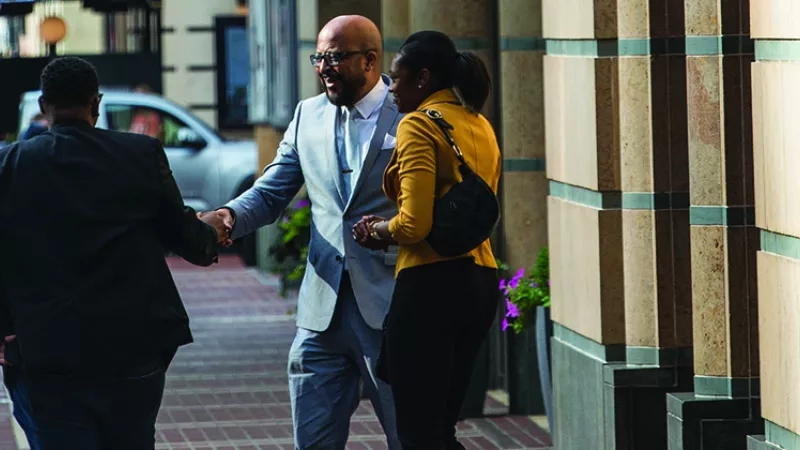 Two men and a woman shake hands outside of a building with a car in the background.