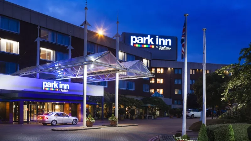 The exterior of Park Inn Hotel at night with a parked car, flags, and blooming flowers.