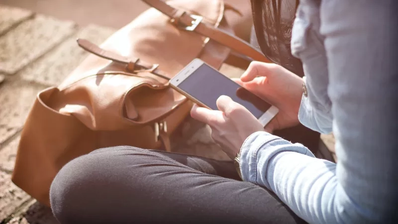 A woman uses her smartphone outside in the sunlight next to a brown leather bag.