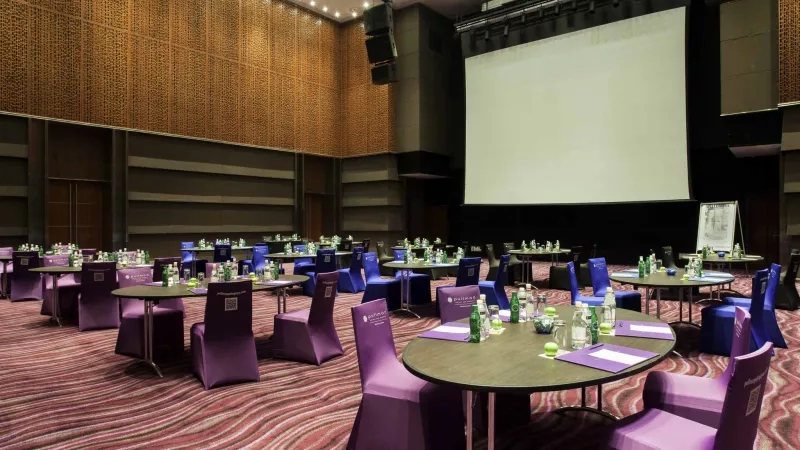 Meeting room at Pullman Putrajaya Lakeside with round tables, purple chairs, and a large screen.