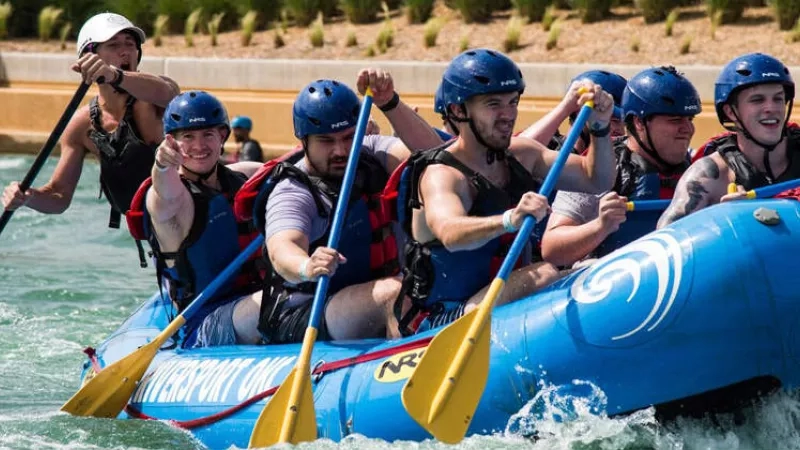 Group of men on a blue raft in the water with paddles and blue helmets.