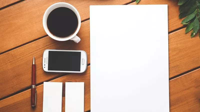 A paper, a cellphone, a cup of coffee, a pen, and two business cards on a wooden table.