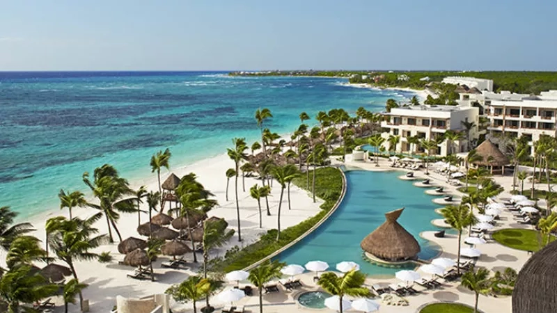 Panoramic view of pool and beach with huts, lounge chairs, and palm trees at Secrets Maroma Beach Riviera Cancun.