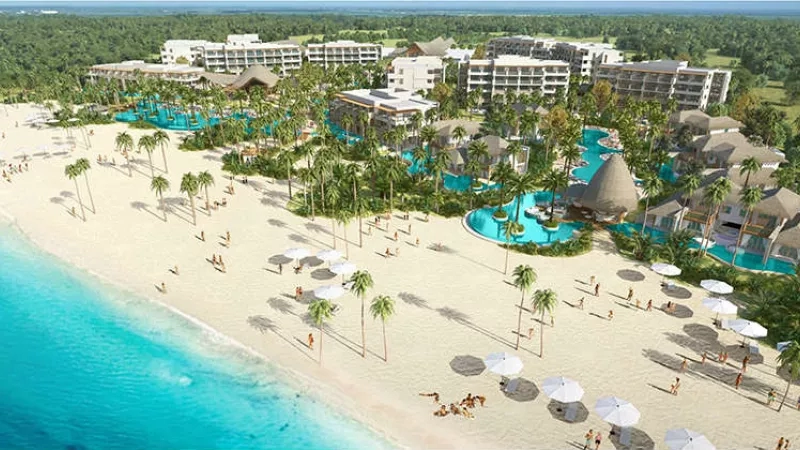 An aerial view of the beachfront resort with pool, buildings, trees, and beach with people and umbrellas.