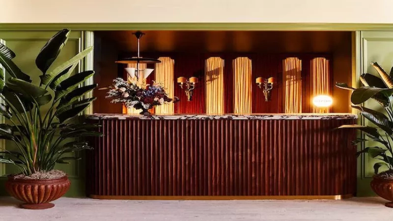 A hotel reception with a wooden desk, green walls, and potted plants on either side.