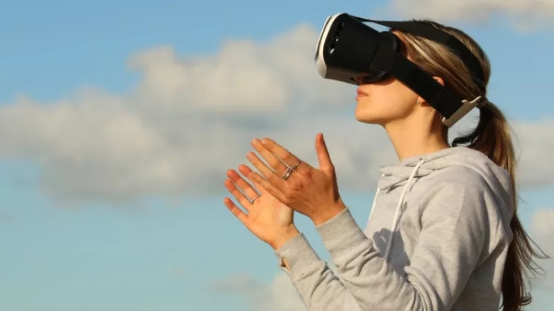Woman wearing a VR headset and clapping her hands in front of a blue sky with clouds.