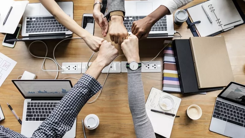 Overhead shot of colleagues holding hands with laptops, notebooks, and other office equipment on a table.