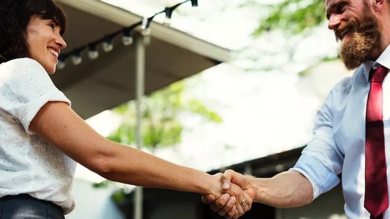 A bearded man and a woman shake hands outside a house with string lights and greenery.