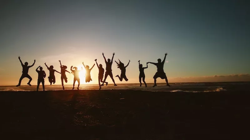 A group of people jumping in the air on a beach at sunset with their arms raised.