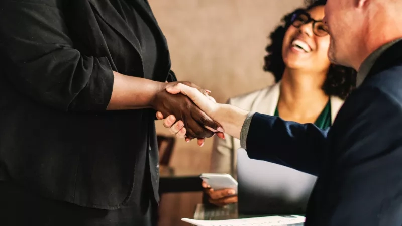 Two people shake hands in a cafe as a woman behind them smiles and looks at her phone.