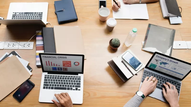 Adults sitting around a table with laptops open, one screen showing "Financial Crisis" and the other "Company's Growth."