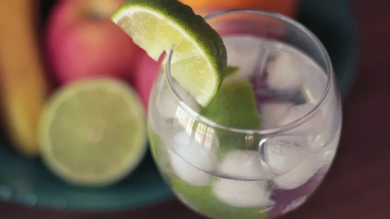 Glass of cocktail with lime slice and ice cubes with fruits on the background in a restaurant bar.
