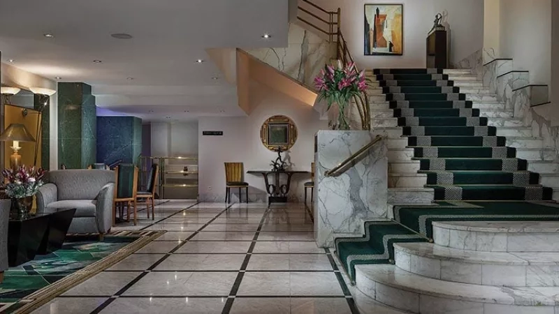 The Alcron Hotel lobby with carpeted stairs, marble pillars, and a flower vase on the landing.