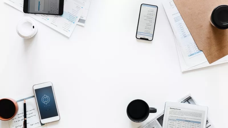 A desk with phones, tablets, papers, coffee, and other items on a white background.