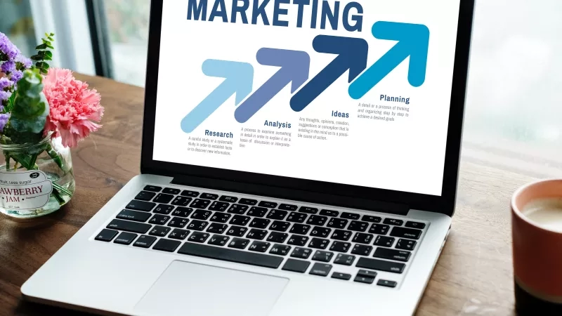 Laptop displaying marketing flowchart with planning, ideas, analysis, and research arrows on a wooden table.