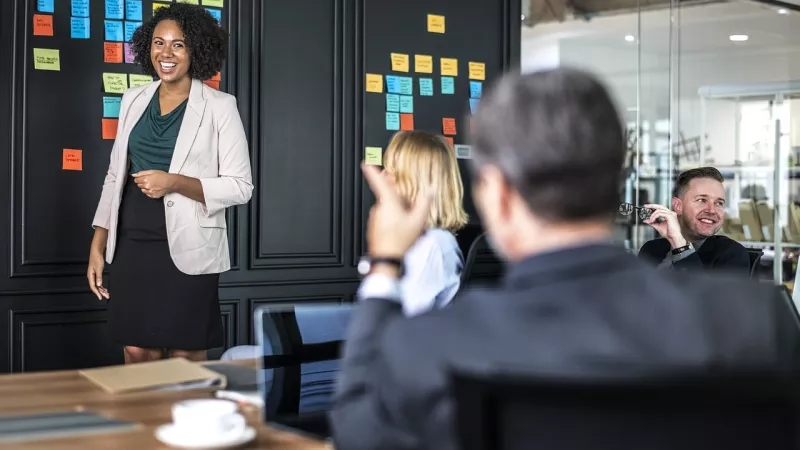 A group of people are in a meeting with sticky notes on the wall and a woman giving a presentation.