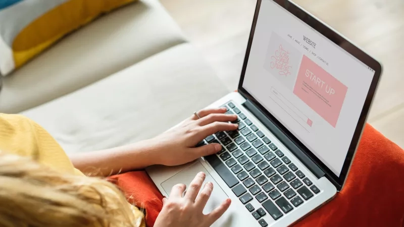 A woman is sitting on a couch using a laptop with a website displayed on the screen.