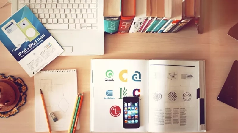 A desk with an Apple iPhone, books, pencils, and an open laptop on it.