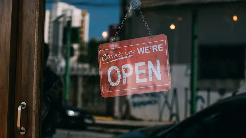 Close up of a red open sign hanging in a business window with cars in the background.