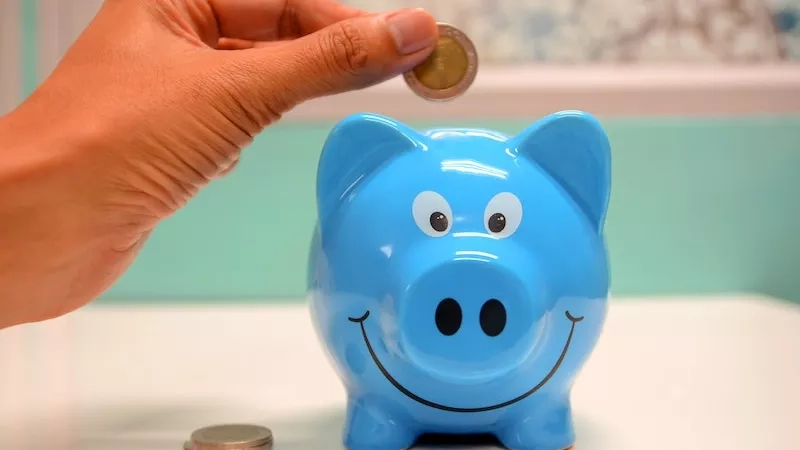Hand placing coin into blue piggy bank in front of pastel-colored wall and mint-colored table.