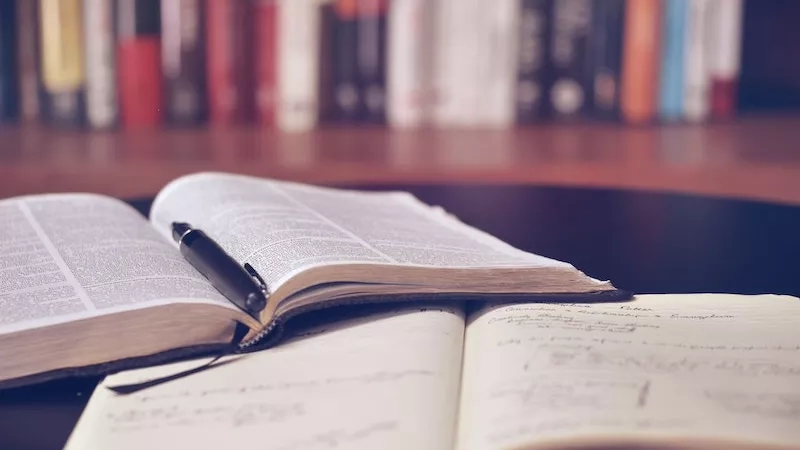 Two open books and pen on table in front of bookshelf with blurred background.