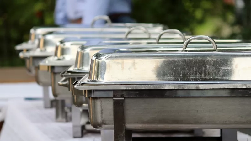 Multiple stainless steel chafing dishes with handles and lids are arranged on a table with a white tablecloth.