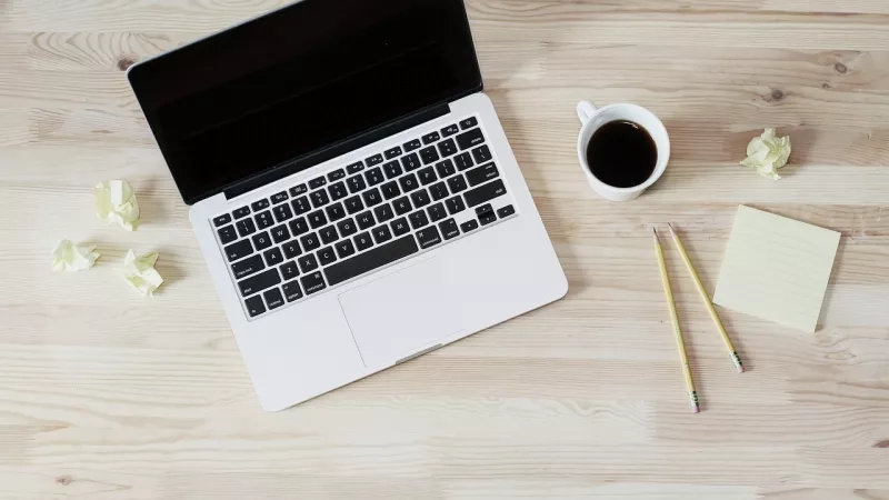 White laptop and coffee cup on a wooden desk with two pencils and three sticky notes.