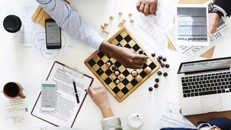 A group of people are playing chess, using tablets and smartphones, and there are papers on the table.