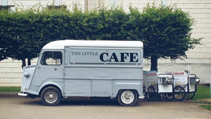 White vintage truck with "The Little Cafe" text parked near trees and a brick building.