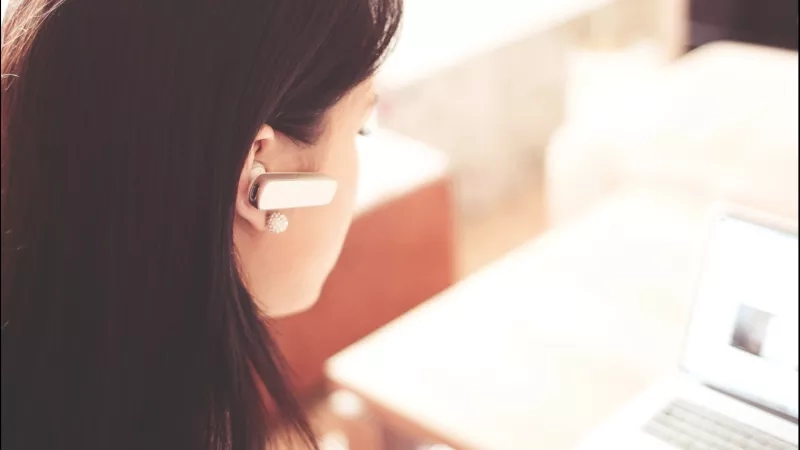 A woman wearing a headset is working on a laptop in a blurred office setting.