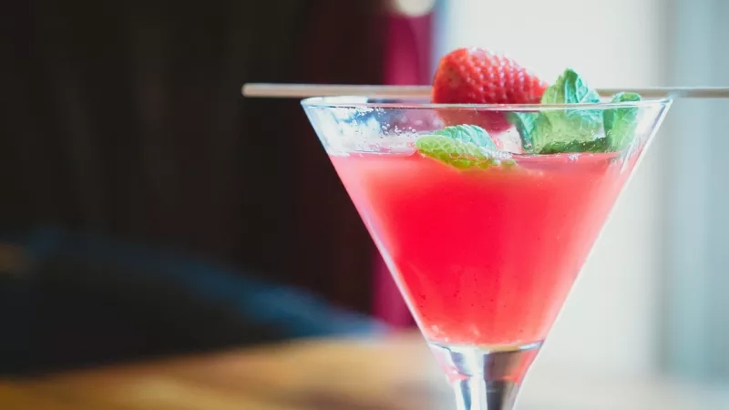 A glass of cocktail with a strawberry and mint leaf garnish and a wooden straw.
