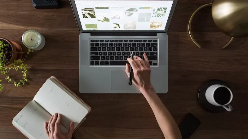 A woman working at a desk with a laptop, notebook, and coffee on a wooden desk.