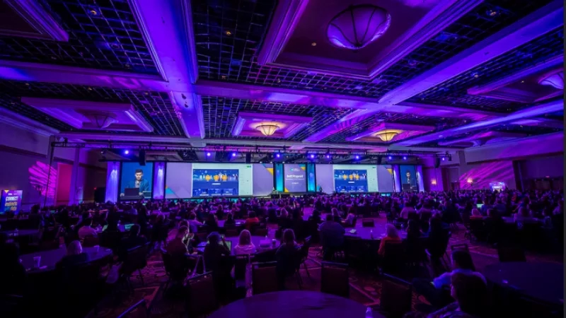 Crowd of people attending a conference with purple lights illuminating the room and three screens on the wall.