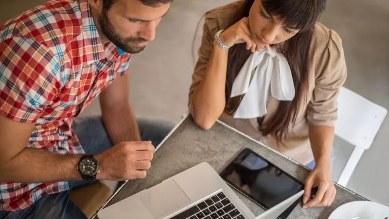 Man and woman looking at laptop and tablet with food on the table in front of them.