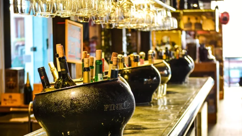 Corked bottles of wine in black buckets on a counter with wine glasses hanging above.
