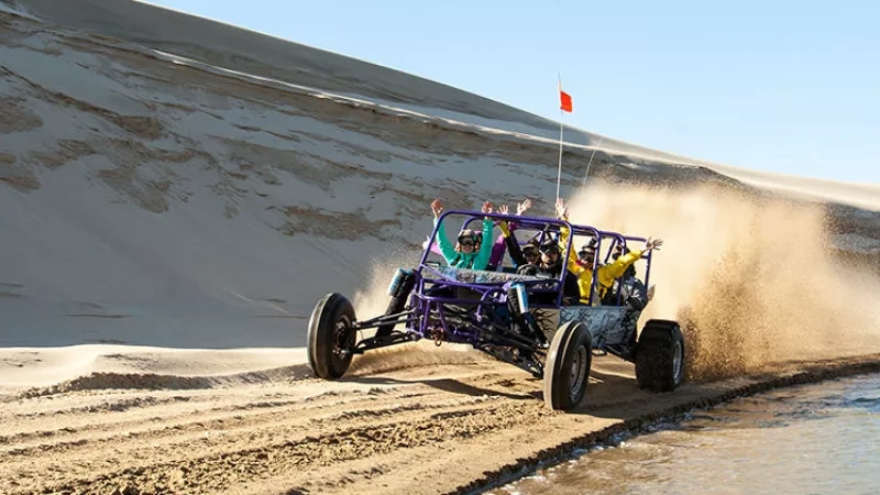 A group of people in a dune buggy on a sandy dune with a flag in the background.