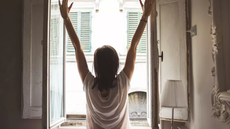 Woman standing at window with arms up in the air, stretching with a smile on her face.