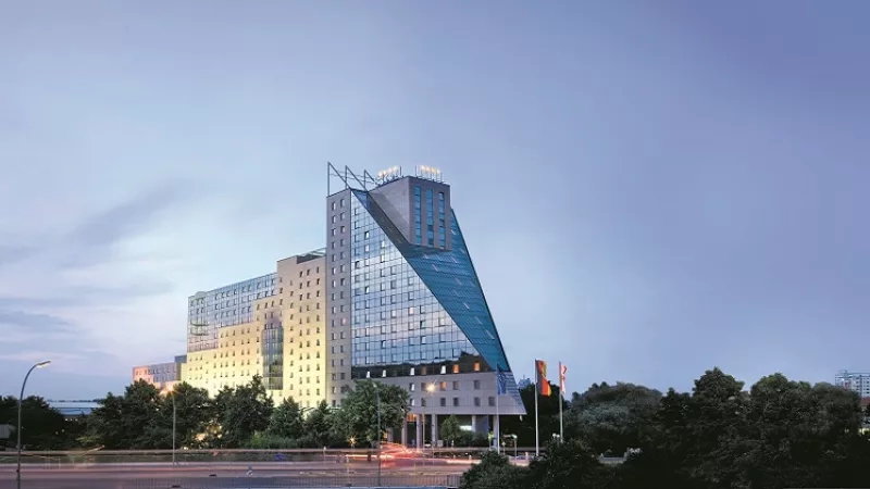 Dusk view of the Estrel Berlin hotel building, with trees and street lights in the foreground.