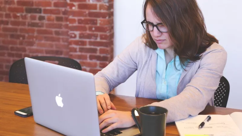 A woman is working on a laptop with an open notebook in front of a brick wall.