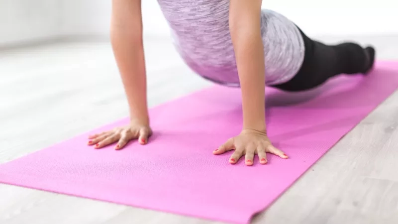 A woman doing push-ups on a pink yoga mat on a wooden floor with white walls.