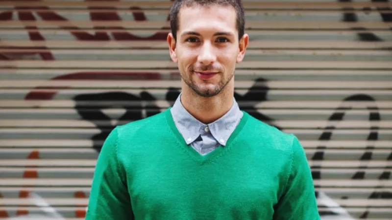 A man wearing a green sweater in front of a wall with graffiti on it.