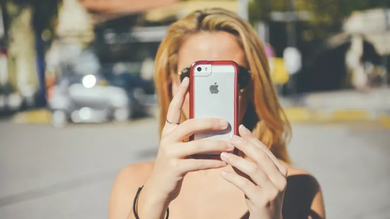 A woman is taking a selfie with an Apple phone while standing on a road with cars.