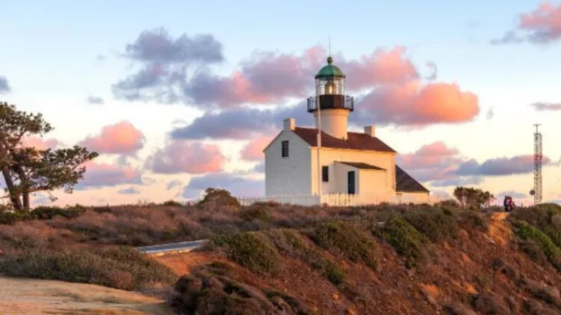 San Diego lighthouse with sunset sky, surrounded by greenery and visitors on the cliff.