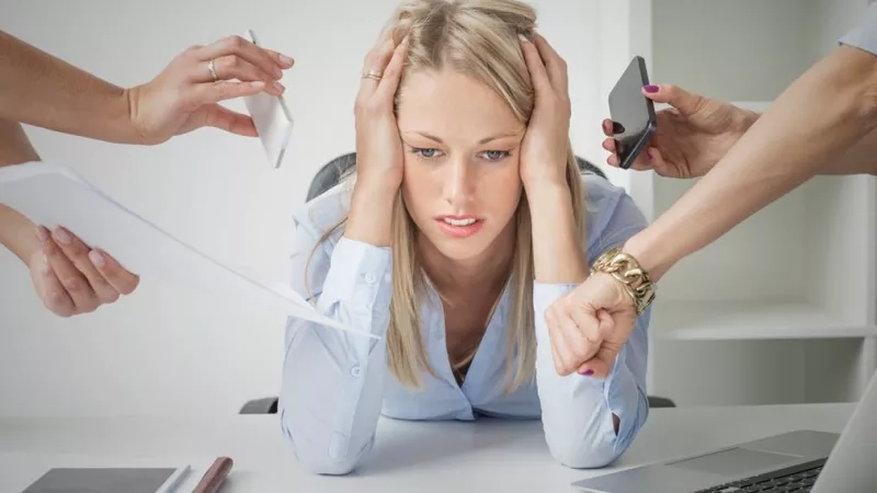 Stressed woman sitting at desk in office with colleagues surrounding her and putting pressure on her to perform.