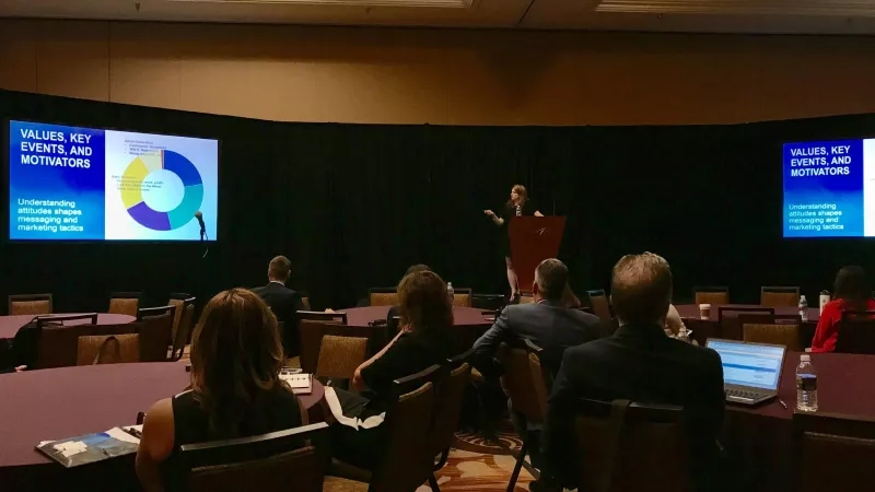 A conference room with a podium, a speaker, and attendees, with a screen displaying a pie chart.
