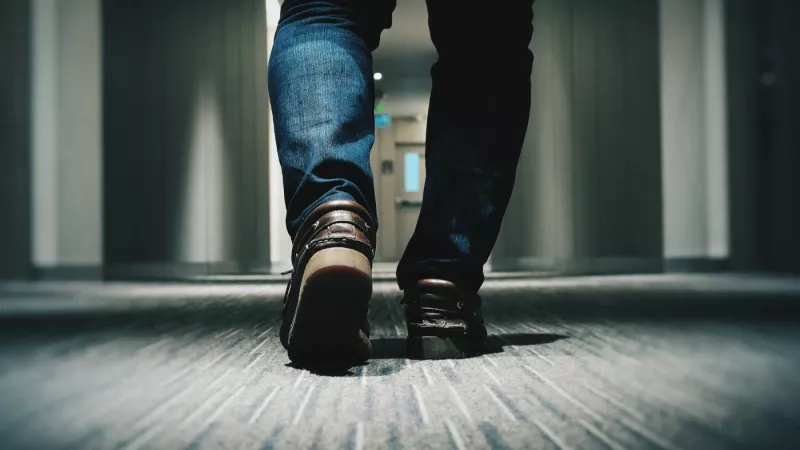 Feet walking down a hotel corridor with light shining on the floor and dark background.