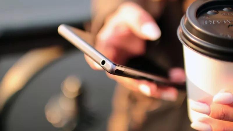 Close up of hands holding a smartphone and coffee with a blurred background.