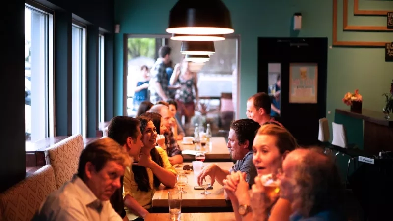 Group of people sitting at tables in a restaurant with blue walls and light fixtures above them.